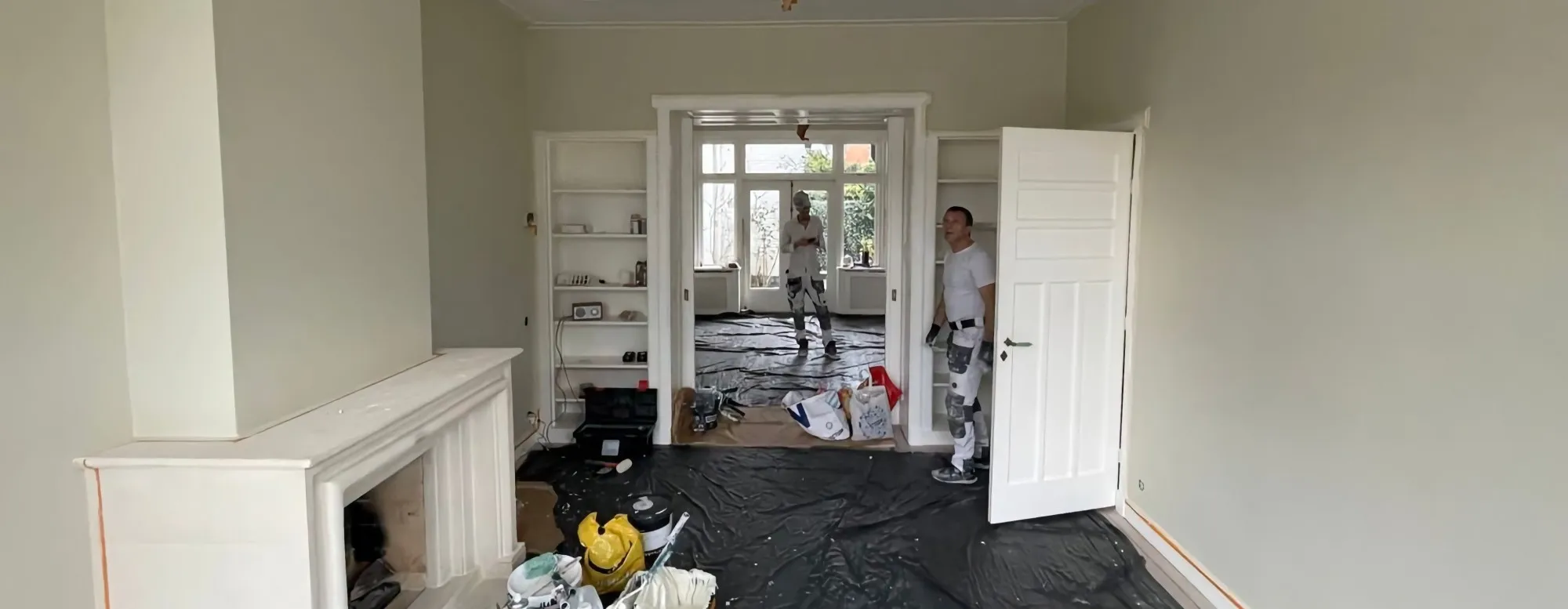 Twee schilders van Flying Design in een woonkamer met afgedekte vloer, witte deuren en schouw